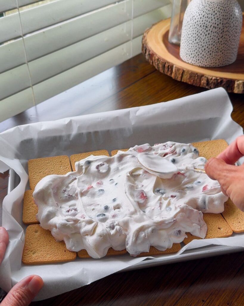 Graham crackers being layered with Greek yogurt, whipped cream, and berries before freezing into sandwich snacks