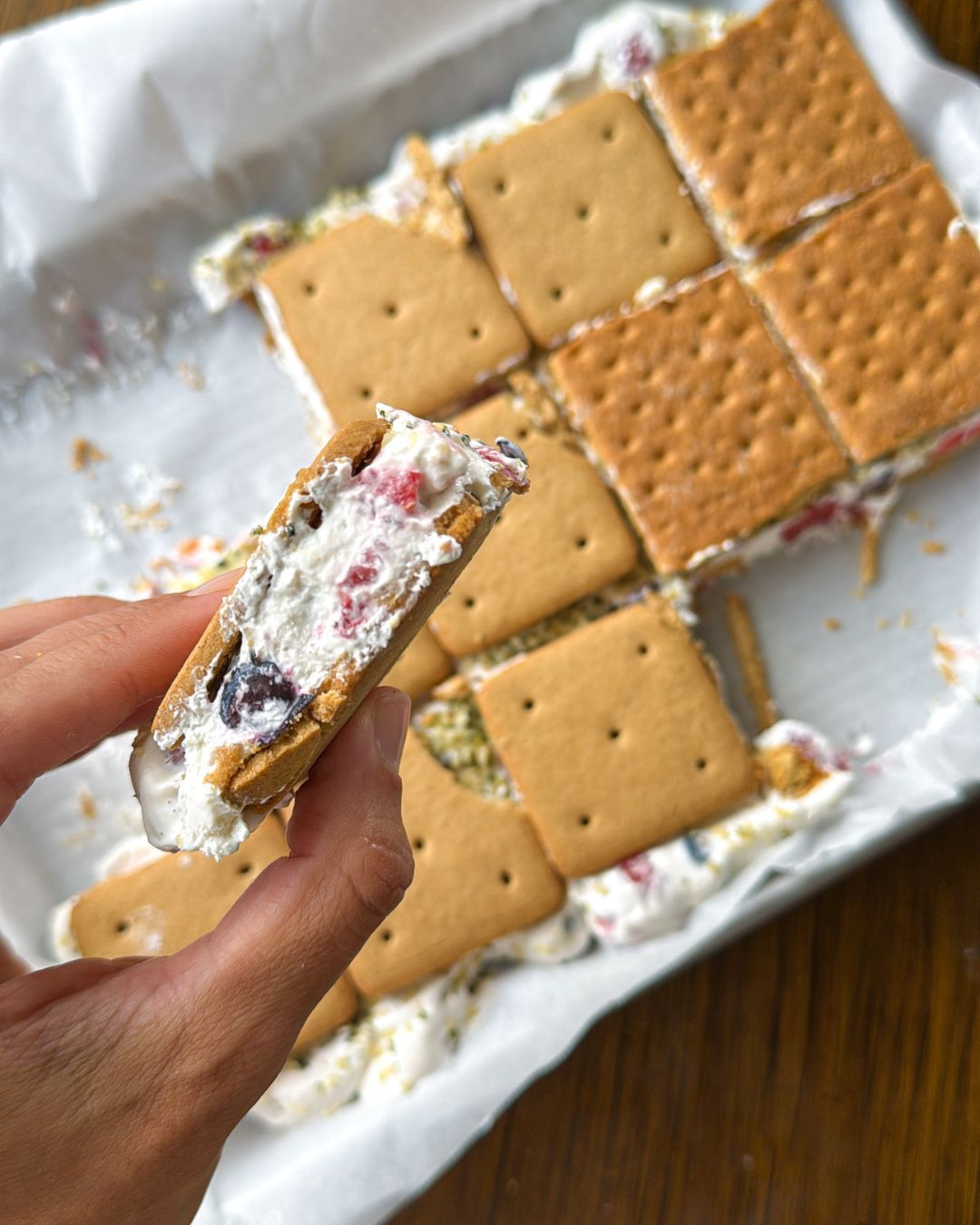 Graham crackers being layered with Greek yogurt, whipped cream, and berries before freezing into sandwich snacks